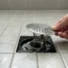 Hand lifting a shower drain cover to reveal a hair and soap scum clog underneath